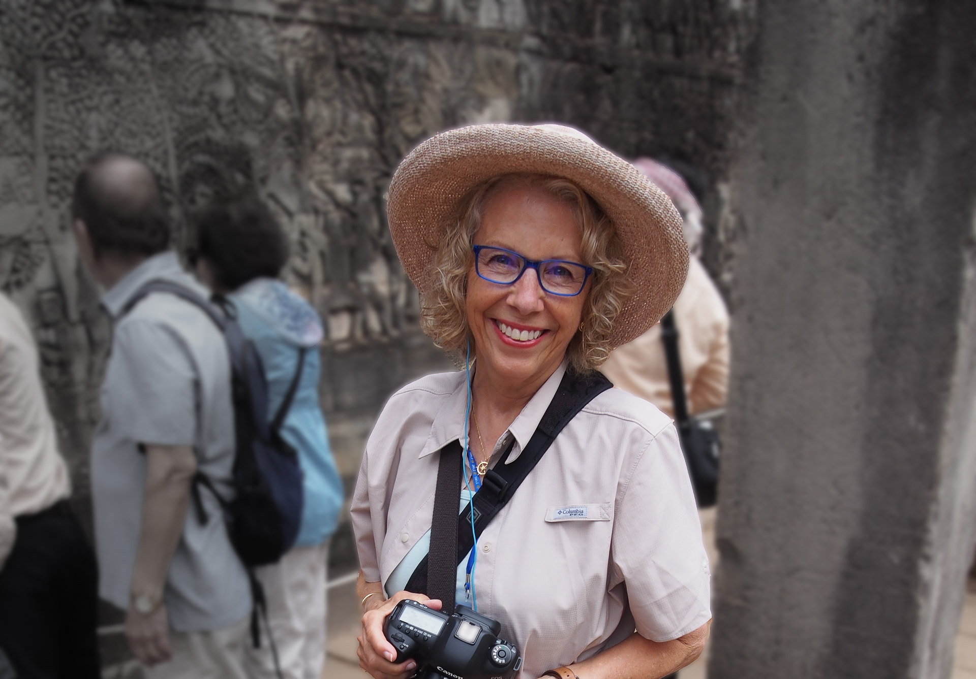 A smiling woman wearing a hat and glasses, holding a camera.