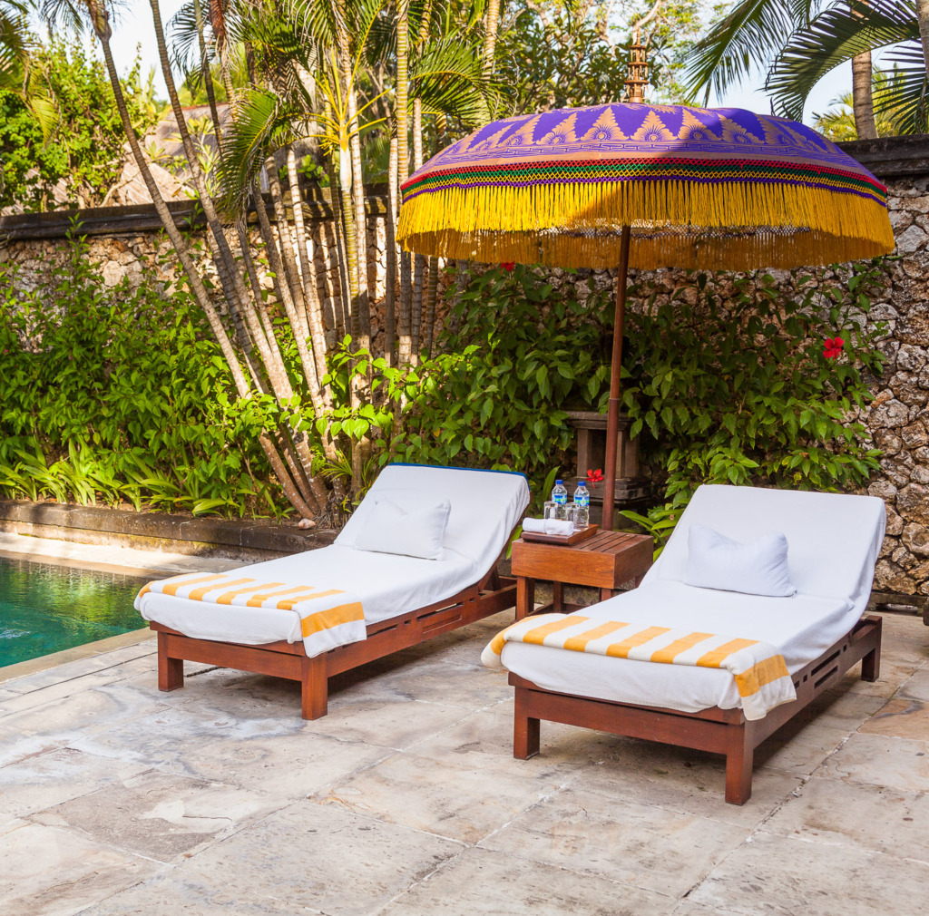Photo of 2 lounge chairs under a purple Indonesian sun umbrella by a swimming pool.