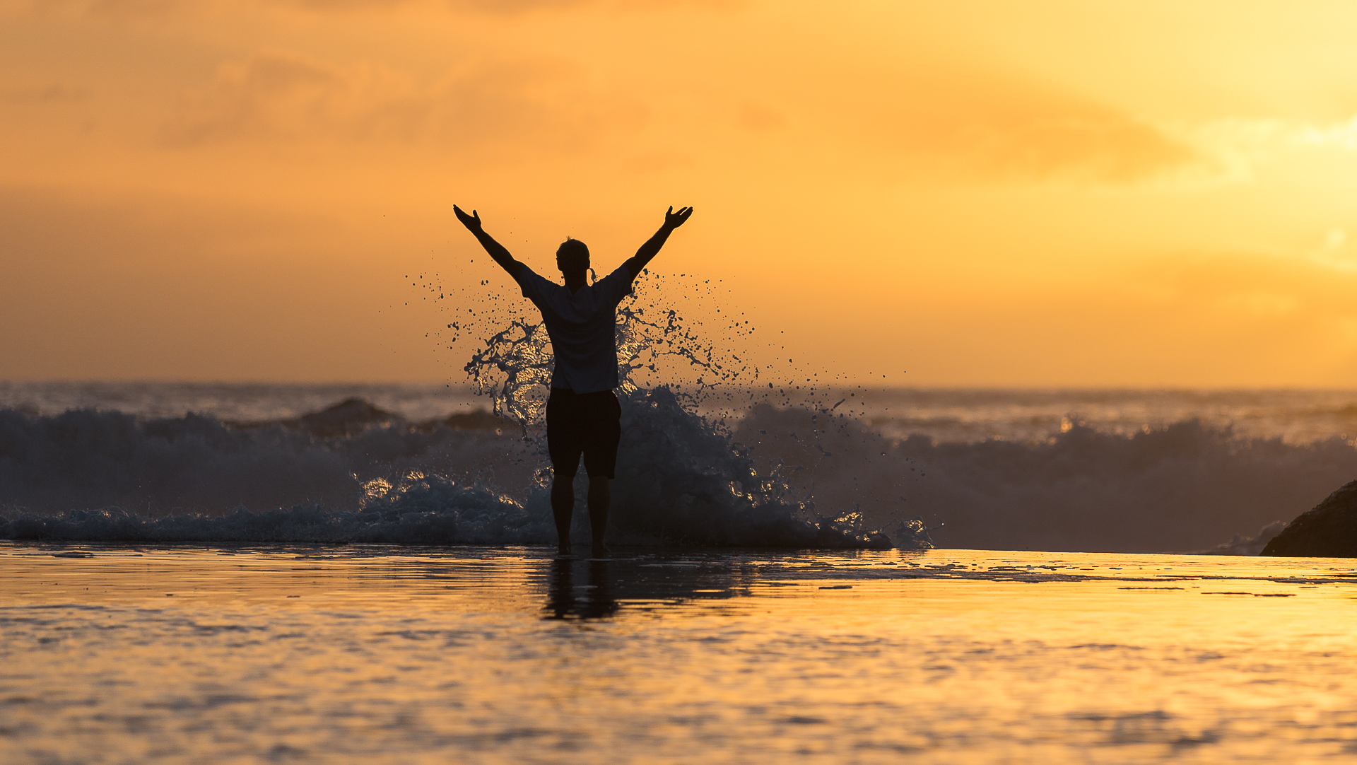 A man lifts his arms and looks at the sunset. He is standing on a sea wall and a wave is breaking against him.