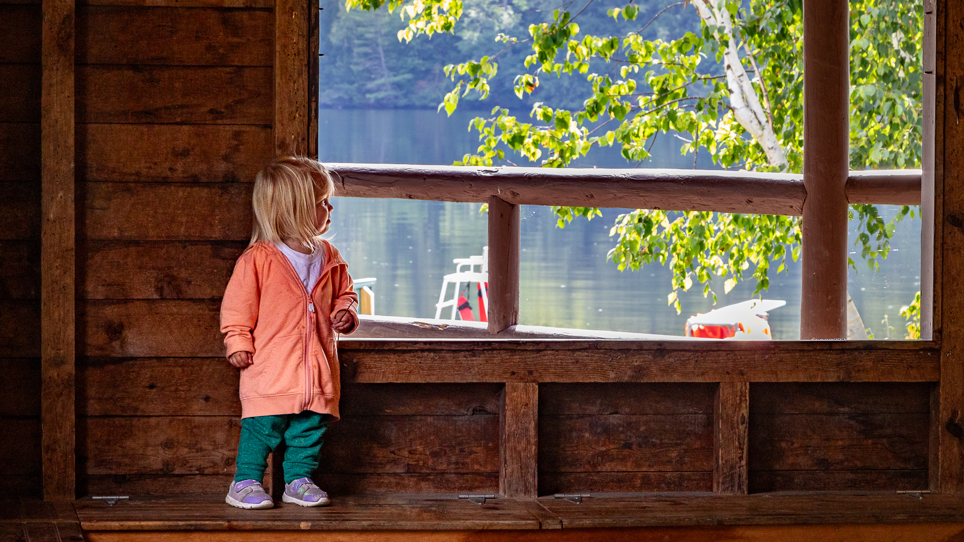 Small child looking out of open cabin window at lake