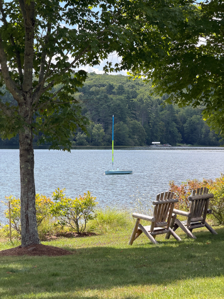 An end-of-summer photo of a two rustic chairs along a lake facing a single sailboat that's tied up. The foliage along the lake is turning orange.