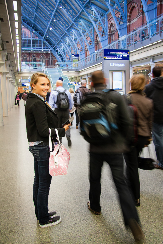 Take better photos indoors by using the correct shutter speed to capture an image of action at St Pancras train station in London like this one of a young woman smiling at camera while other travelers rush past.