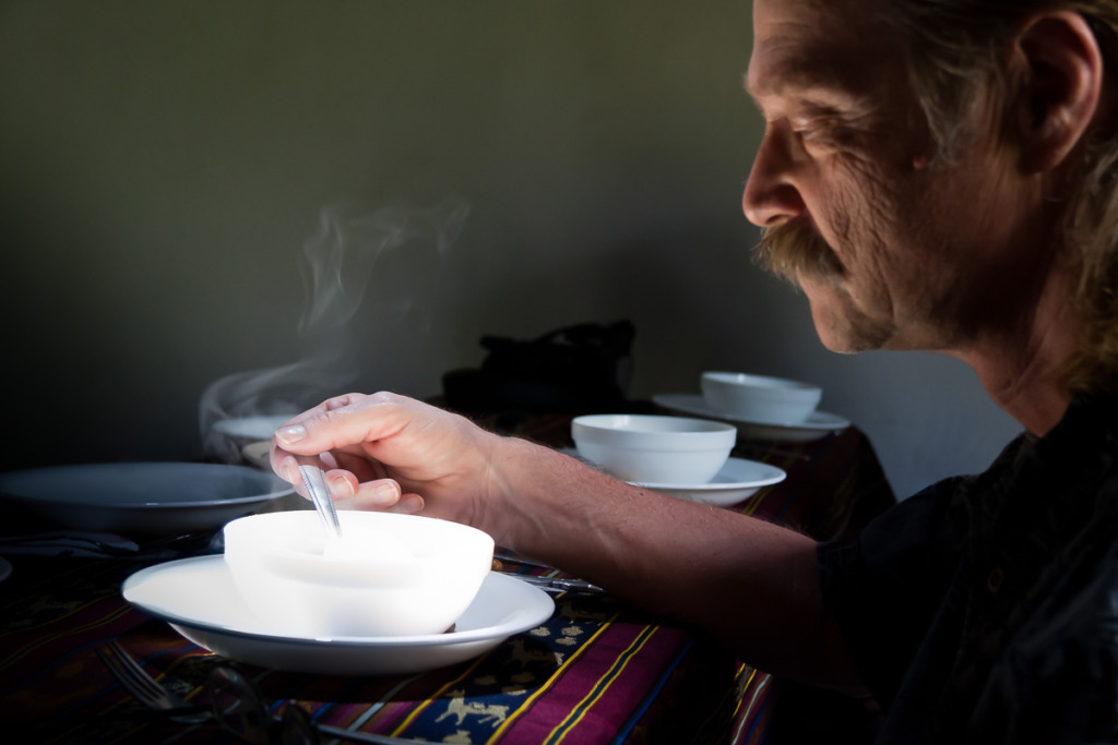 A ray of sun lights a steaming bowl of soup as a man is stirring it.