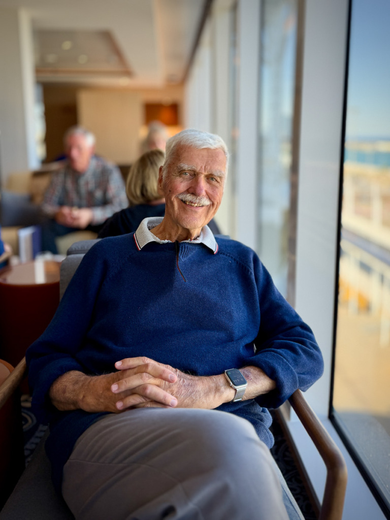 Man in a chair on a cruise ship smiling at camera. The people behind him are out of focus because photographer knew how to use aperture mode.